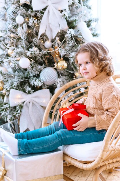 Jolie petite fille heureuse avec des boucles est assise sur une chaise près de l'arbre de Noël avec une boîte-cadeau dans les mains