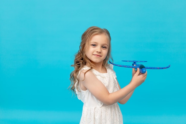 Jolie petite fille heureuse dans une robe blanche en coton sur fond bleu avec un avion dans ses mains jouant une place pour le texte