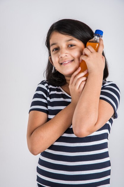 Jolie petite fille indienne ou asiatique avec une bouteille pour animaux de compagnie de boisson froide à l'orange ou à la mangue ou de jus de fruits, buvant ou tenant debout isolé sur fond bleu ou blanc.