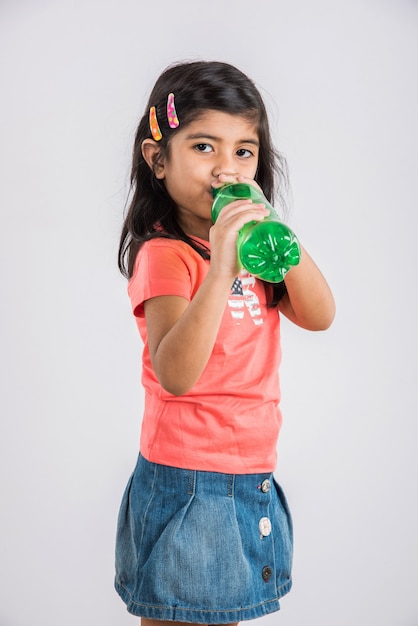 Jolie petite fille indienne ou asiatique avec une bouteille pour animaux de compagnie de boisson froide à l'orange ou à la mangue ou de jus de fruits, buvant ou tenant debout isolé sur fond bleu ou blanc.