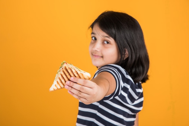 Jolie petite fille indienne ou asiatique mangeant un délicieux hamburger, un sandwich ou une pizza dans une assiette ou une boîte. Debout isolé sur fond bleu ou jaune.