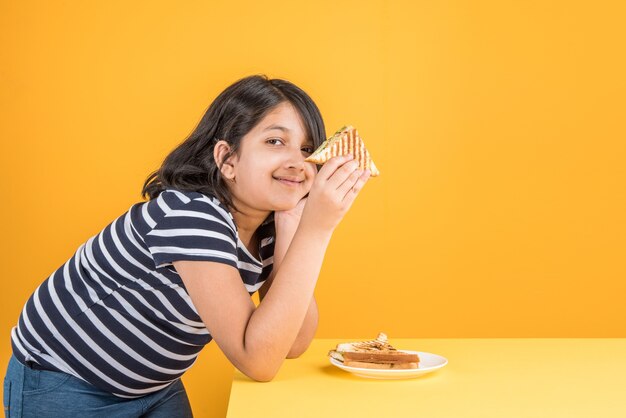 Jolie petite fille indienne ou asiatique mangeant un délicieux hamburger, un sandwich ou une pizza dans une assiette ou une boîte. Debout isolé sur fond bleu ou jaune.