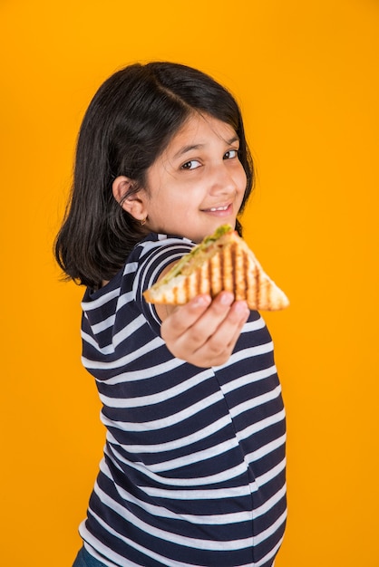 Jolie petite fille indienne ou asiatique mangeant un délicieux hamburger, un sandwich ou une pizza dans une assiette ou une boîte. Debout isolé sur fond bleu ou jaune.