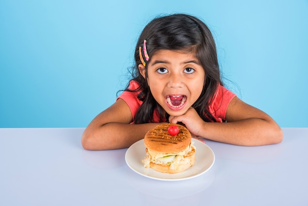 Jolie petite fille indienne ou asiatique mangeant un savoureux hamburger, sandwich ou pizza dans une assiette ou une boîte. Debout isolé sur fond bleu ou jaune.