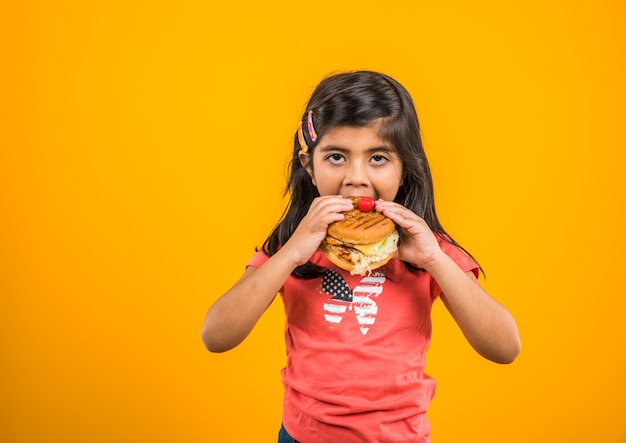 Jolie petite fille indienne ou asiatique mangeant un savoureux hamburger, sandwich ou pizza dans une assiette ou une boîte. Debout isolé sur fond bleu ou jaune.