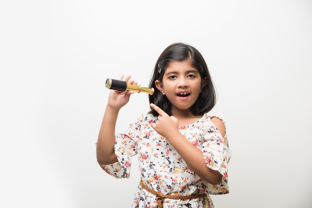 Jolie petite fille indienne ou asiatique utilisant un télescope extensible et regardant dans le ciel, veut être une scientifique. Debout isolé sur fond de tableau blanc ou vert