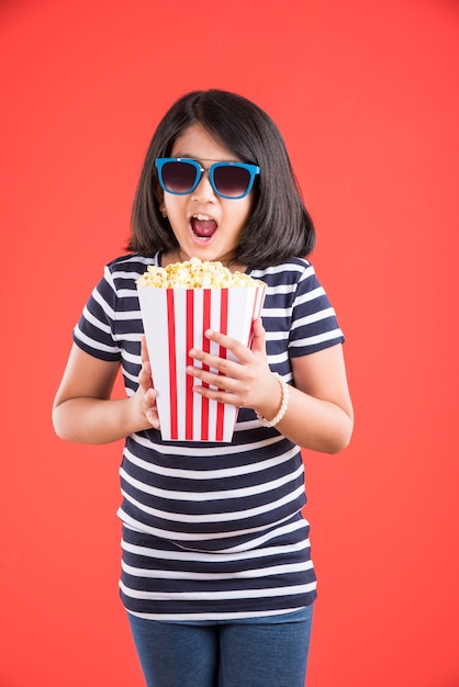 Jolie petite fille indienne mangeant du pop-corn tout en portant des lunettes de soleil ou des lunettes 3D dans un théâtre. Debout isolé sur fond coloré