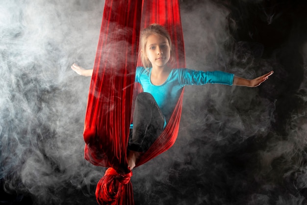 Jolie petite fille intrépide dans un costume de gymnastique bleu montre un ruban rouge aérien stunt entouré de fumée sur un fond noir