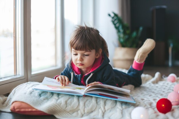 Jolie petite fille lisant un livre allongé sur une couverture en laine près de la fenêtre. c'est l'hiver, il neige et il fait froid dehors.