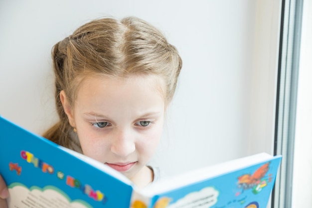 Jolie petite fille lisant un livre sur le rebord de la fenêtreportrait d'une belle petite fille européenne se concentrant...