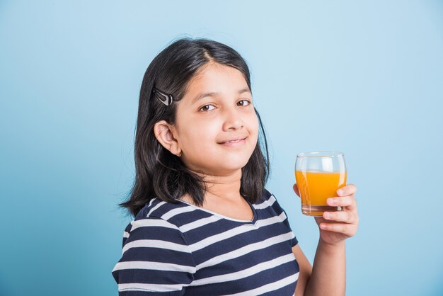 Jolie petite fille ludique indienne ou asiatique buvant de la mangue fraîche ou du jus d'orange ou une boisson froide ou une boisson dans un verre, isolée sur fond blanc