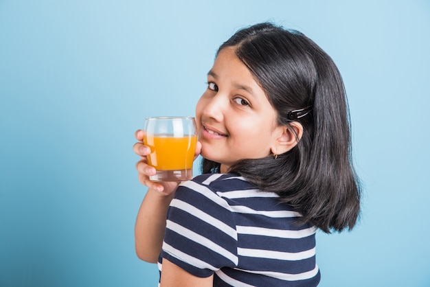 Jolie petite fille ludique indienne ou asiatique buvant de la mangue fraîche ou du jus d'orange ou une boisson froide ou une boisson dans un verre, isolée sur fond blanc