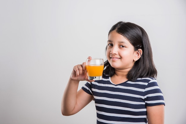 Jolie petite fille ludique indienne ou asiatique buvant de la mangue fraîche ou du jus d'orange ou une boisson froide ou une boisson dans un verre, isolée sur fond blanc