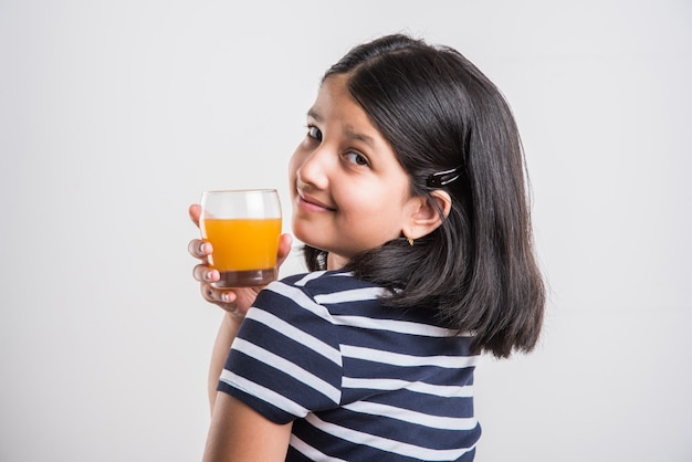 Jolie petite fille ludique indienne ou asiatique buvant de la mangue fraîche ou du jus d'orange ou une boisson froide ou une boisson dans un verre, isolée sur fond blanc