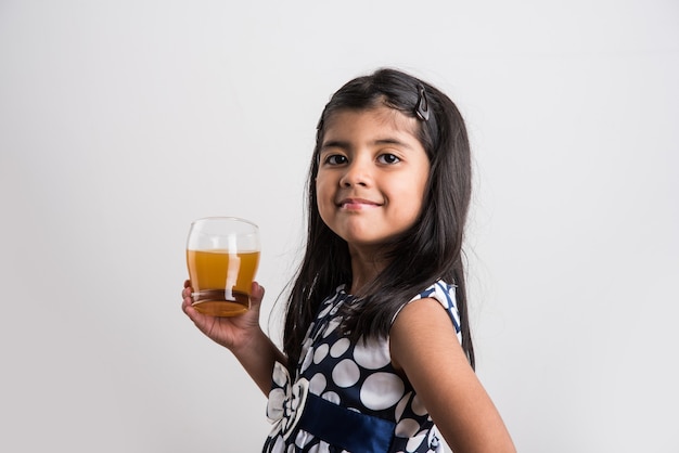 Jolie petite fille ludique indienne ou asiatique buvant de la mangue fraîche ou du jus d'orange ou une boisson froide ou une boisson dans un verre, isolée sur fond blanc