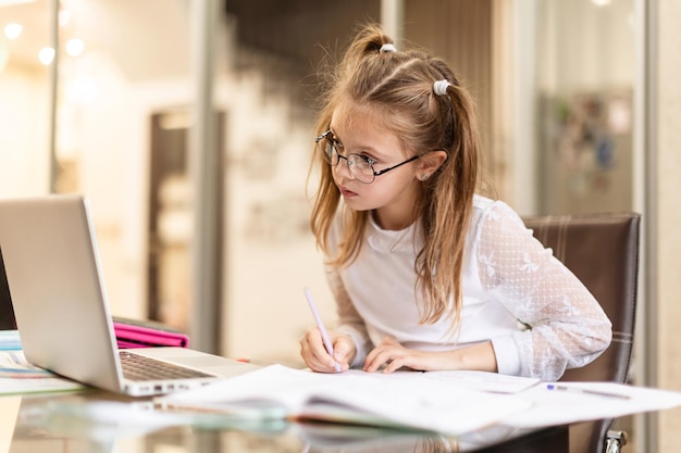 Jolie petite fille à lunettes faisant sérieusement ses devoirs à l'aide d'un ordinateur portable à la maison pour l'éducation en ligne, l'étude à domicile. Enseignement à distance pour les enfants. Distance sociale. Reste à la maison.