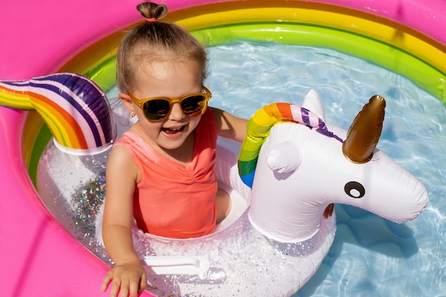 une jolie petite fille à lunettes de soleil nage avec une bouée de sauvetage en forme de licorne dans une piscine gonflable