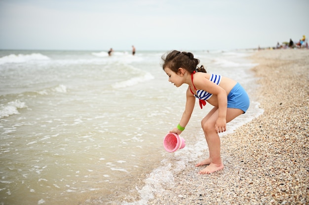 Jolie petite fille en maillot de bain recueillant de l'eau de mer dans un seau de jouet rose Enfant mignon jouant sur th