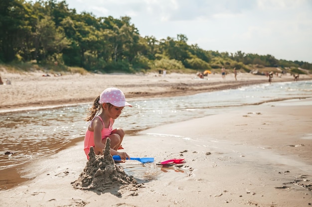 Jolie petite fille en maillot de bain rose jouant avec du sable et de l'eau sur la plage d'une mer Baltique