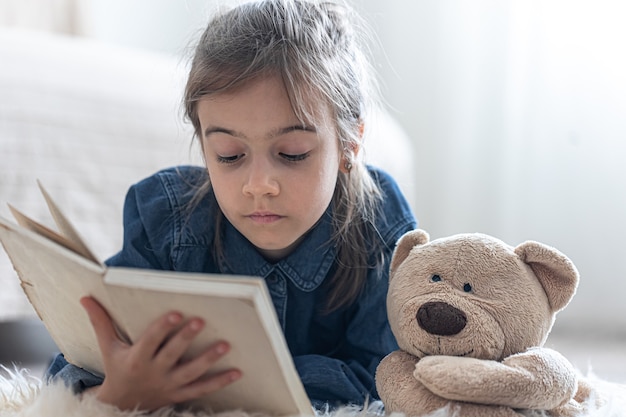 Jolie petite fille à la maison, allongée sur le sol avec son jouet préféré et lit un livre.