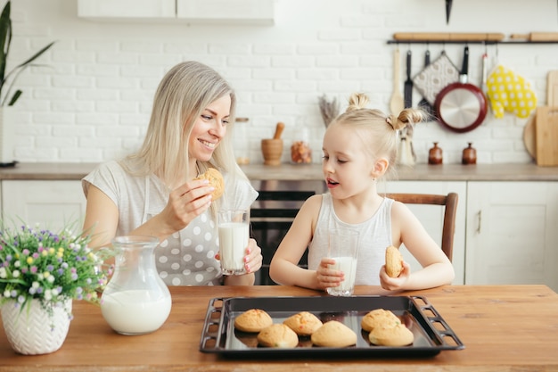 Jolie petite fille et maman mangeant des biscuits fraîchement préparés avec du lait dans la cuisine. Famille heureuse. Tonifiant.