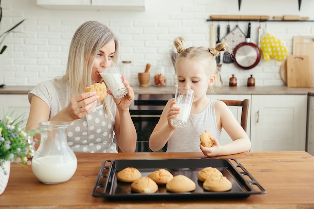 Jolie petite fille et maman mangeant des biscuits fraîchement préparés avec du lait dans la cuisine. Famille heureuse. Tonifiant.