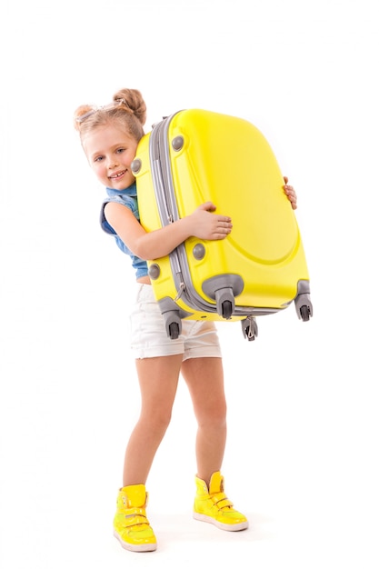 Jolie petite fille mignonne en chemise bleue, short blanc et lunettes de soleil tiennent une valise jaune