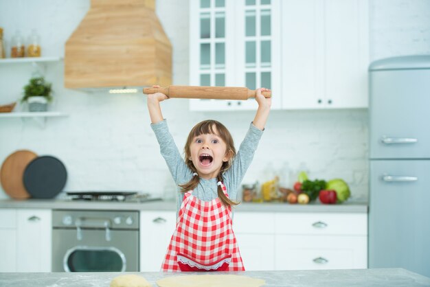 Une jolie petite fille avec des nattes dans un tablier à carreaux pétrit seule la pâte à pizza. Cours de cuisine pour enfants. Émotion de joie.