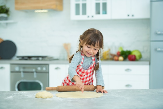 Une jolie petite fille avec des nattes dans un tablier à carreaux pétrit seule la pâte à pizza. Cours de cuisine pour enfants.