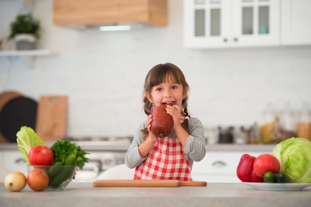 Jolie petite fille avec des nattes dans un tablier culinaire à carreaux mangeant des saucisses dans la cuisine à table.