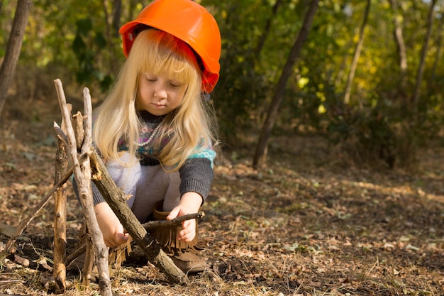 Jolie petite fille ouvrier du bâtiment aux longs cheveux aveugles portant un casque orange accroupi sur le sol construisant un tipi à partir de brindilles de bois