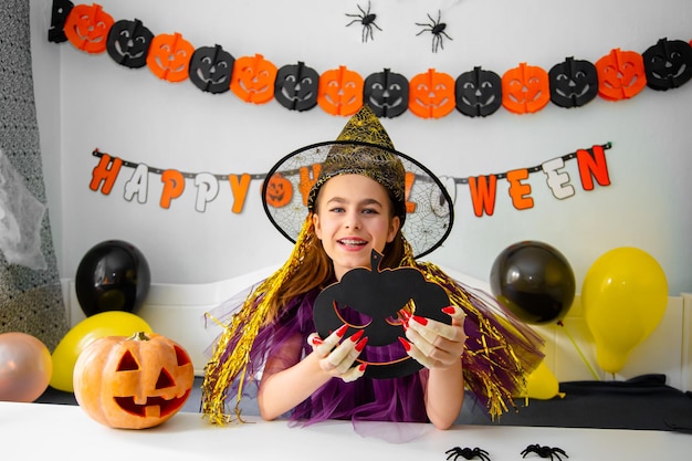 Jolie petite fille portant un chapeau de sorcière assise derrière une table dans une salle décorée sur le thème d'Halloween Covid19