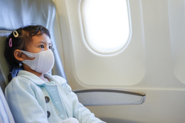 Jolie petite fille portant un masque protecteur pour empêcher les virus assis au siège de la fenêtre dans Airplan