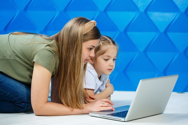 Jolie petite fille prête à utiliser le clavier de l'ordinateur portable assise sur le sol de la cuisine avec sa mère aimante