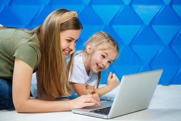 Jolie petite fille prête à utiliser le clavier de l'ordinateur portable assise sur le sol de la cuisine avec sa mère aimante