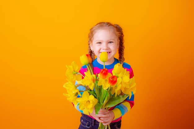 jolie petite fille en pull multicolore tient un bouquet de fleurs printanières sur fond jaune se cachant