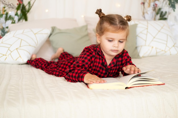 jolie petite fille en pyjama de Noël rouge allongé sur le lit en lisant un livre