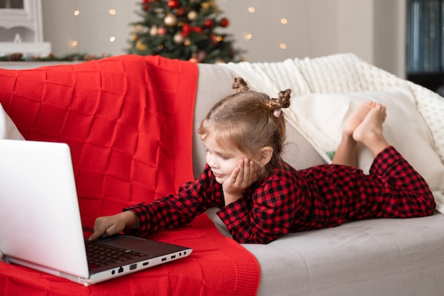 jolie petite fille en pyjama de Noël rouge utilisant un ordinateur portable, ayant un chat vidéo, allongée sur un canapé