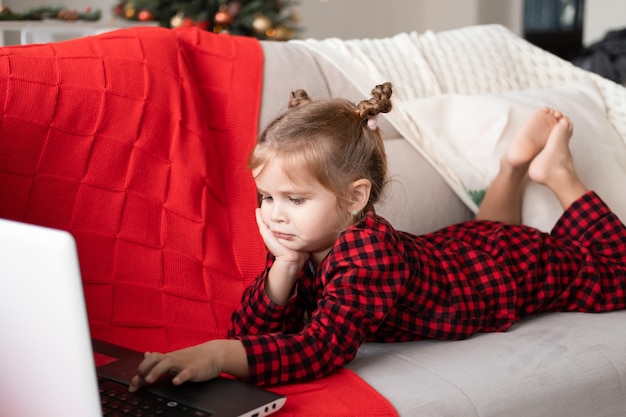 jolie petite fille en pyjama de Noël rouge utilisant un ordinateur portable, ayant un chat vidéo, allongée sur un canapé