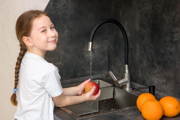 Jolie petite fille avec une queue de cochon est heureuse de laver les fruits dans les pommes et les oranges de la cuisine et sourit