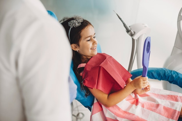 Jolie petite fille regardant dans le miroir ses dents après une chirurgie dentaire dans une stomatologie pédiatrique. Heureux enfant après examen des dents.