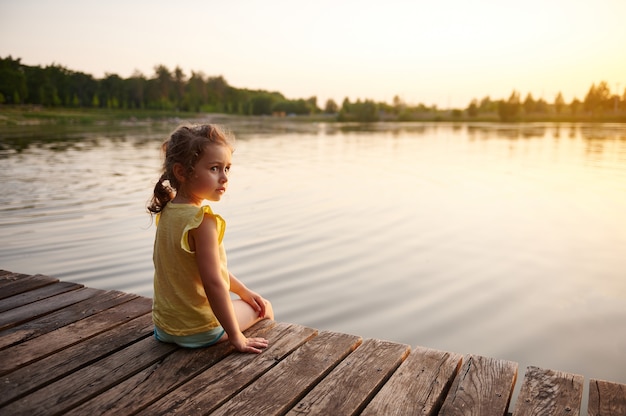 Jolie petite fille reposante sur la jetée au coucher du soleil. Chaude soirée d'été. Chaleur d'été, soirées d'été dans la nature, temps libre au grand air