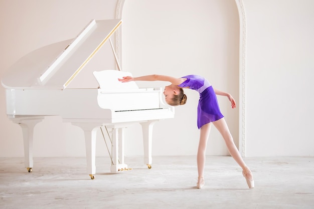 Une Jolie Petite Fille Rêve De Devenir Ballerine Professionnelle Dans Une Salle Blanche à Côté Du Piano, Une Fille En Justaucorps Lilas Danse Sur Des Chaussons De Pointe élève De L'école Professionnelle