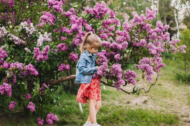 Jolie petite fille en robe riant dans le parc du printemps près de l'enfant lilas sent le lilas