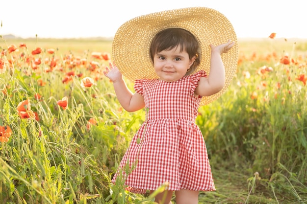 Jolie petite fille en robe rouge et chapeau de paille sur champ de coquelicots au coucher du soleil d'été