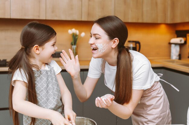 Jolie petite fille et sa jeune mère en tablier jouent avec de la farine et rient en pétrissant la pâte dans la cuisine