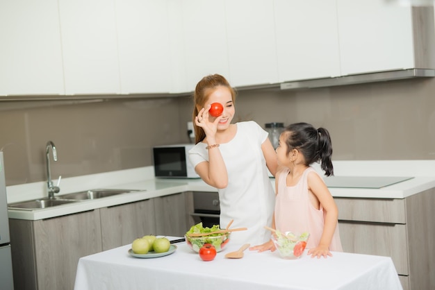 Une jolie petite fille et sa mère en toques coupent des légumes en cuisinant une salade et souriant