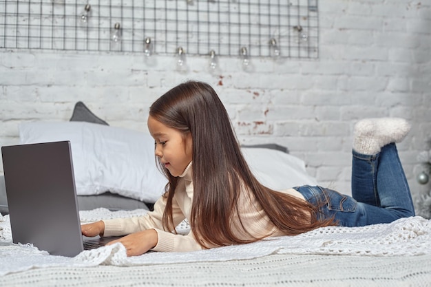 Jolie petite fille se sentant amusante en regardant des dessins animés sur un ordinateur portable assis sur le lit