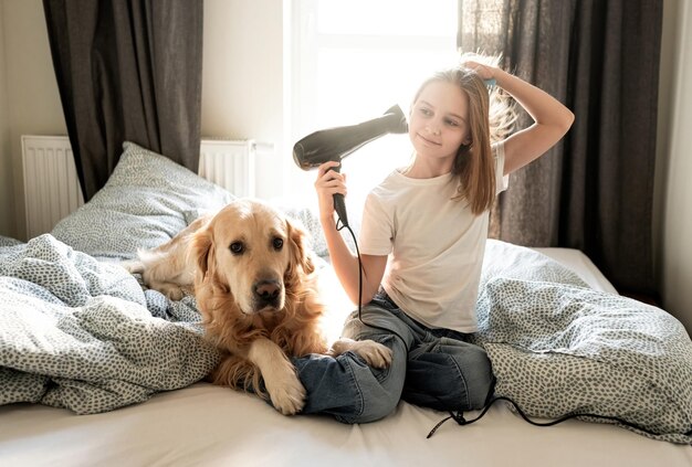 Une jolie petite fille sèche ses cheveux avec un sèche-cheveux assise avec un chien sur un lit.