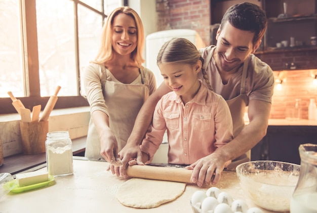 Jolie petite fille et ses beaux parents en tabliers aplatissent la pâte et sourient en cuisinant dans la cuisine à la maison